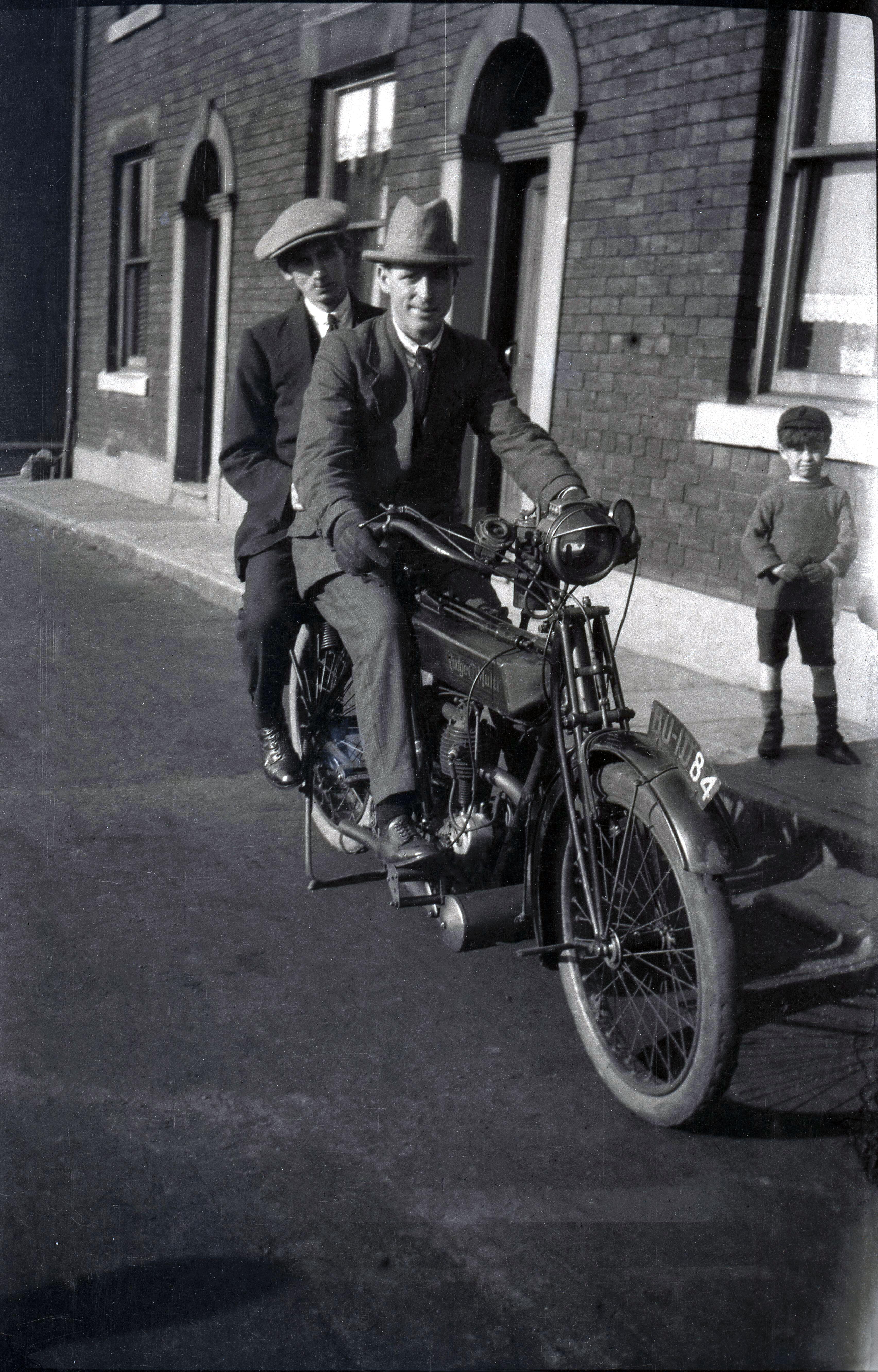Vintage black and white photo of a man on a motorcycle with two children in front of a building.