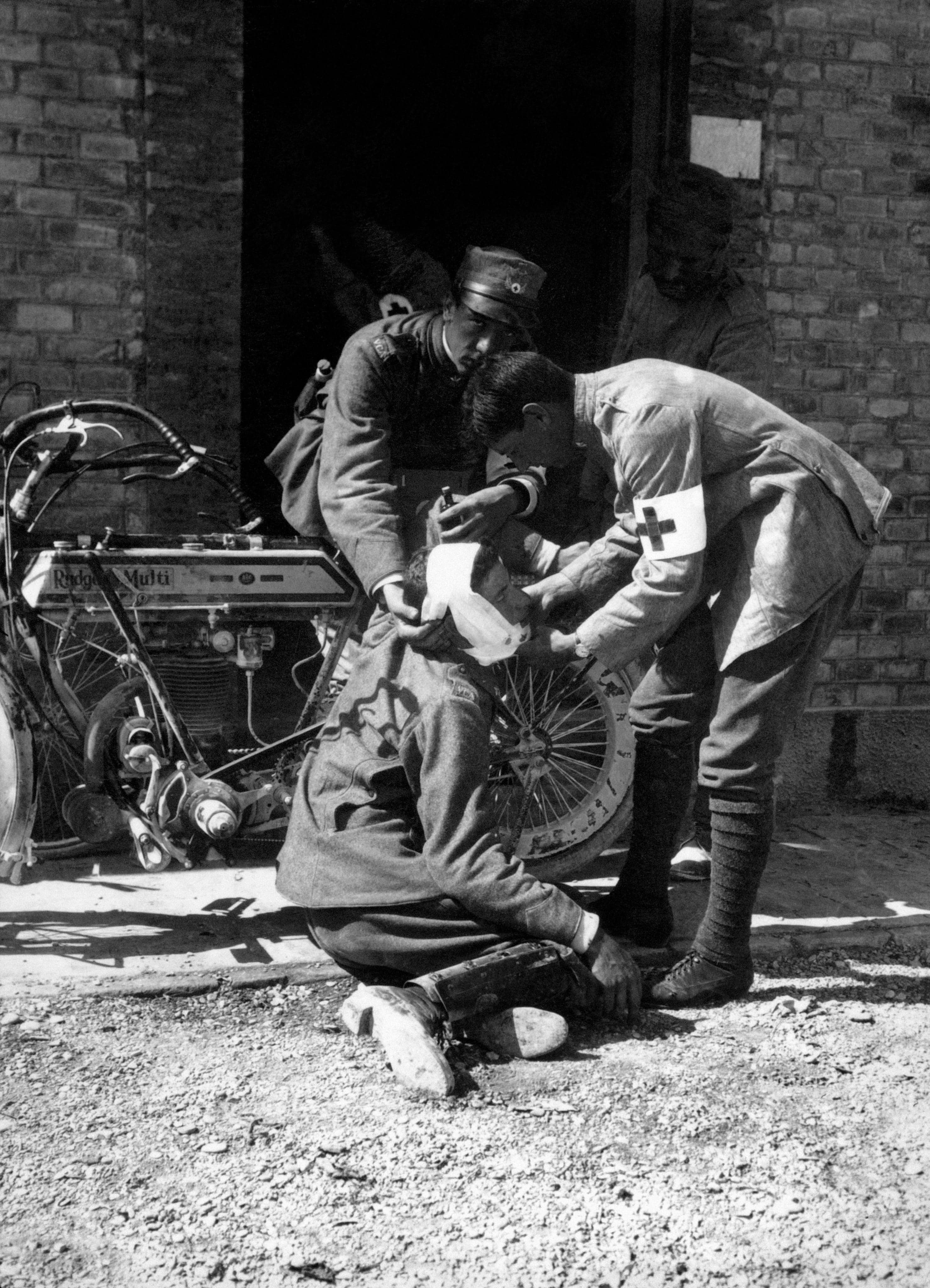 Two soldiers attending to a wounded soldier outdoors, with a brick building in the background.